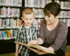 Nainen osoittaa avoinna sylissään olevan kirjan sivua vieressään olevalle pojalle.