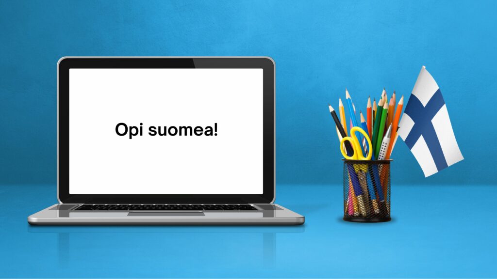 Kannettava tietokone, jonka näytöllä lukee 'Opi suomea!'. Tietokoneen vieressä kynäteline, jossa on värikyniä, sakset ja pieni Suomen lippu. Taustana sininen seinä.